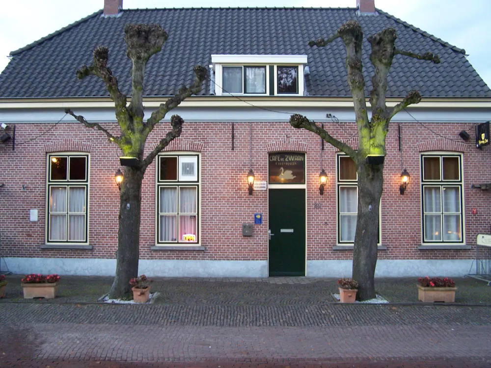 De voorgevel van het café met verlichte lantaarns en twee knotbomen